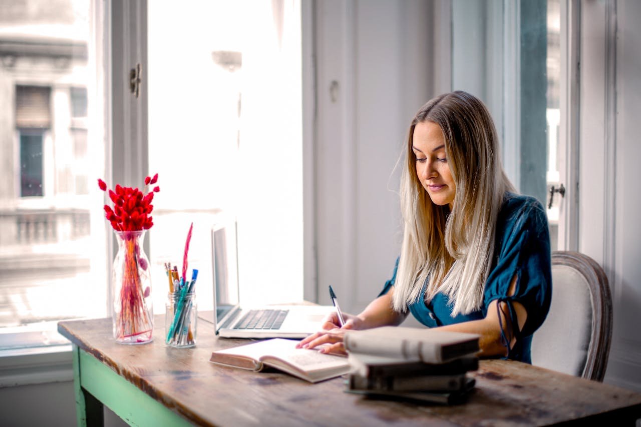 Professional woman working remotely at home office desk, focused on writing and using laptop.