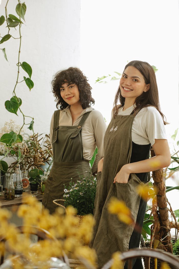 Two cheerful florists in aprons surrounded by lush plants indoors.