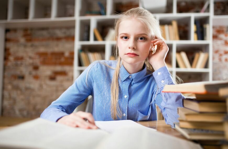 A young woman with blond hair studying at a table piled with books.