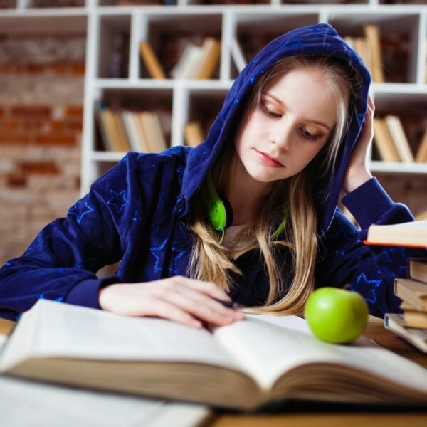 Teenage girl in blue hood reads in library, headphones and apple by her side.