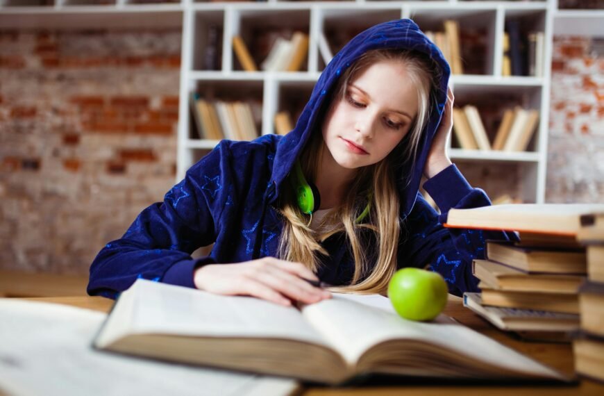 Teenage girl in blue hood reads in library, headphones and apple by her side.