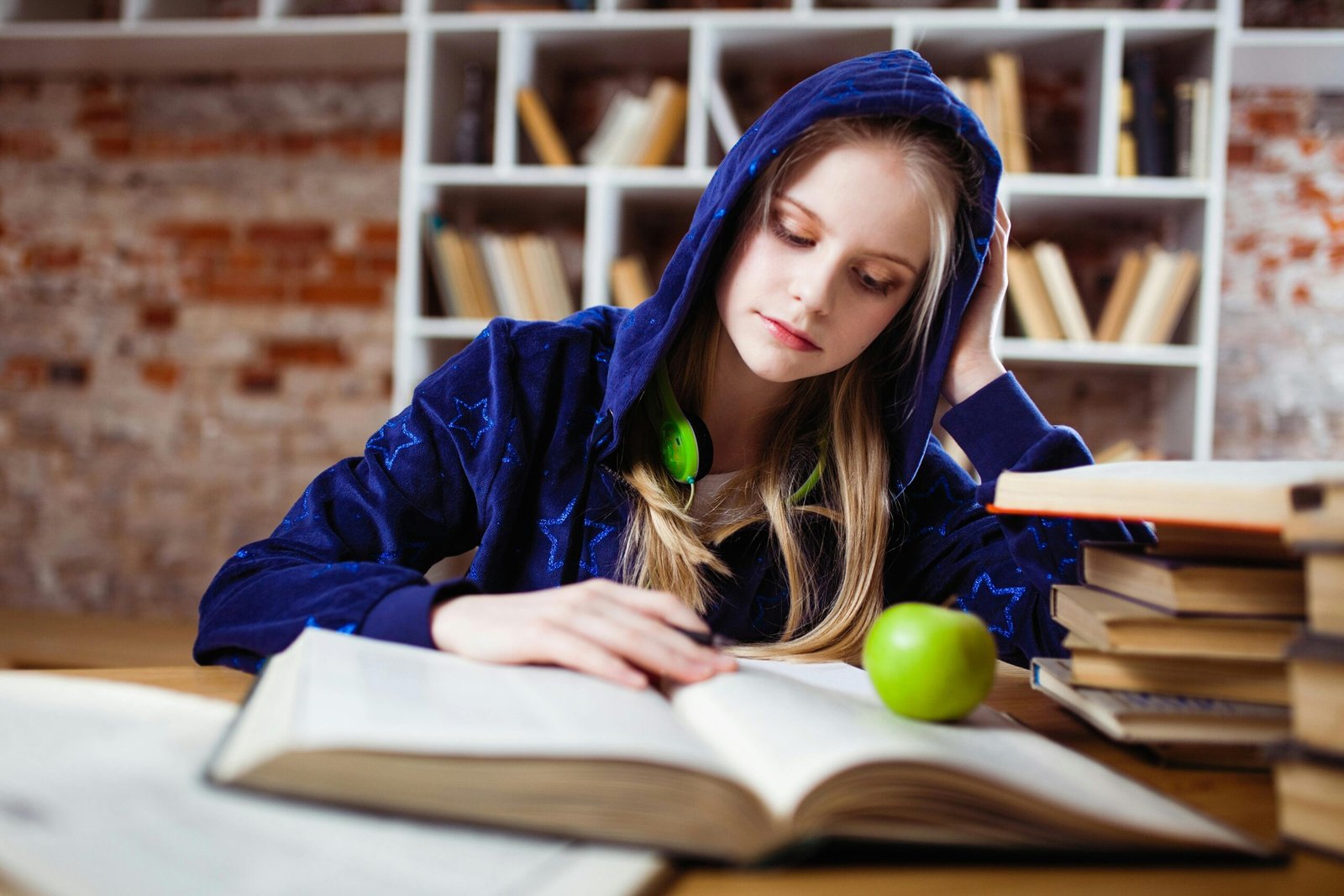 Teenage girl in blue hood reads in library, headphones and apple by her side.