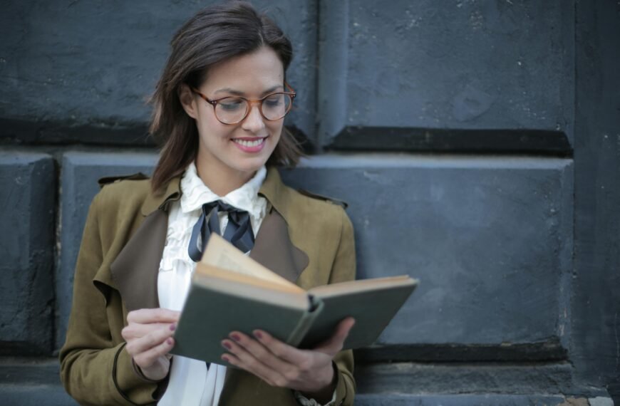 Woman with glasses reading a book against a concrete wall, looking happy and intellectual.