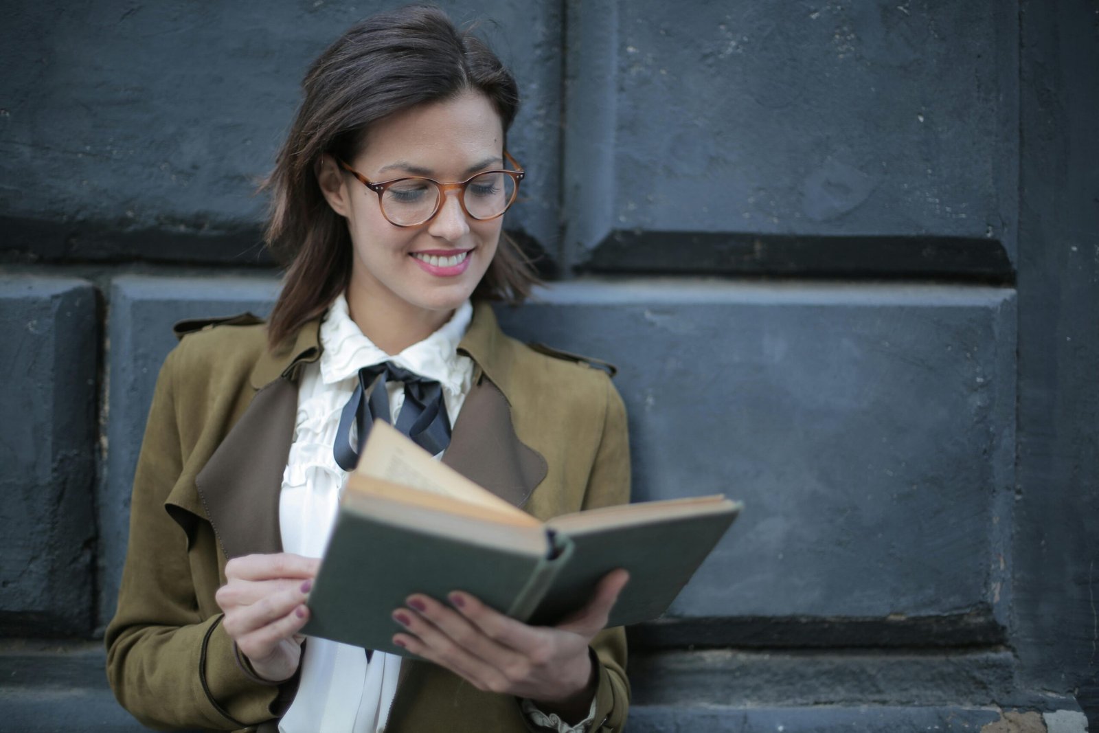 Woman with glasses reading a book against a concrete wall, looking happy and intellectual.