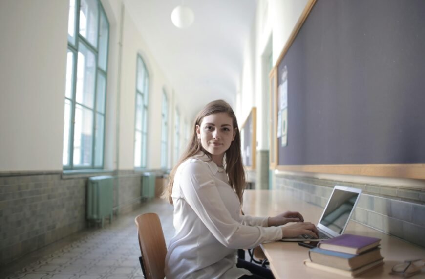 Young woman using laptop in a bright university corridor, seated by a desk, showing focus and concentration.