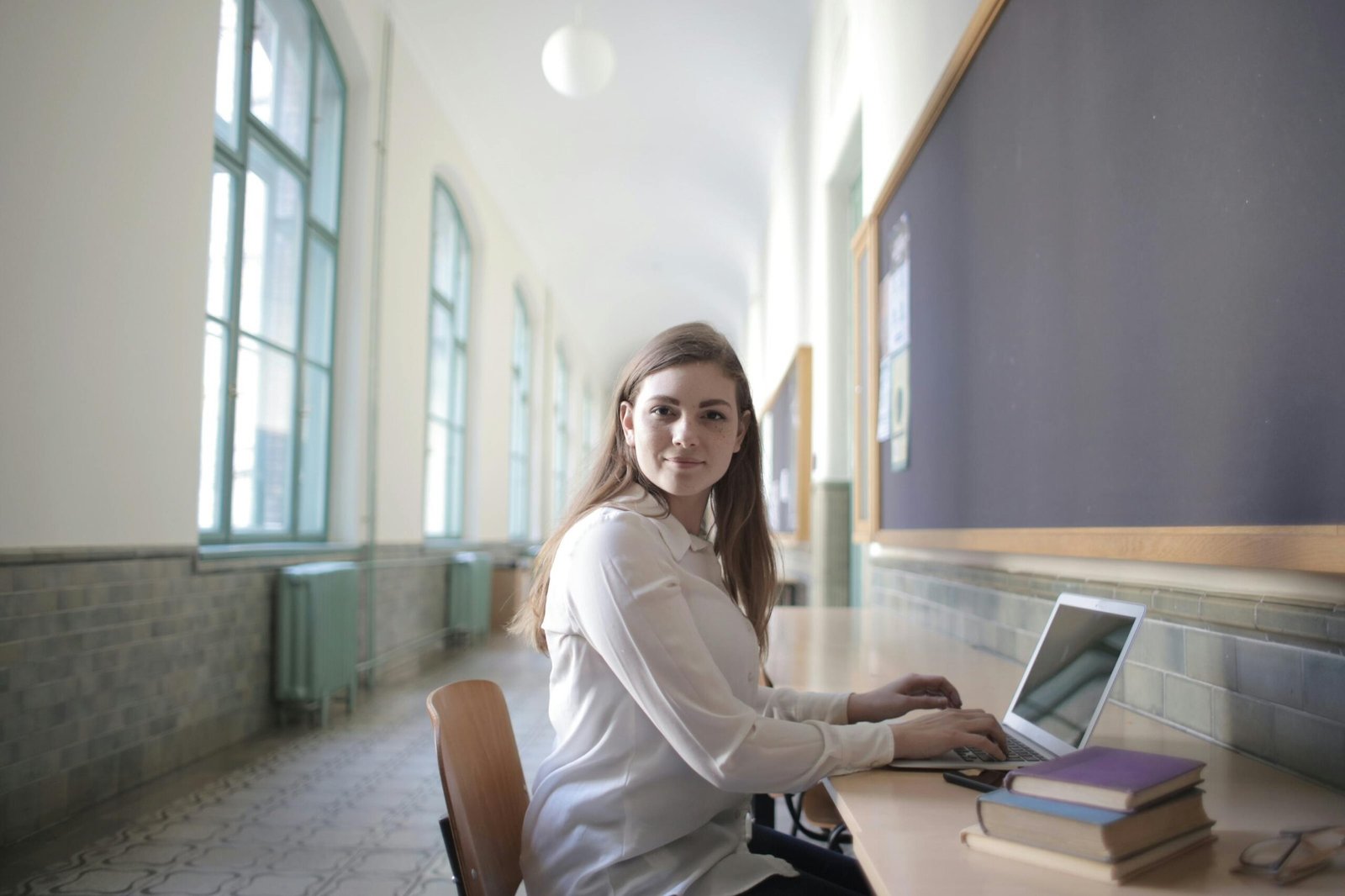 Young woman using laptop in a bright university corridor, seated by a desk, showing focus and concentration.