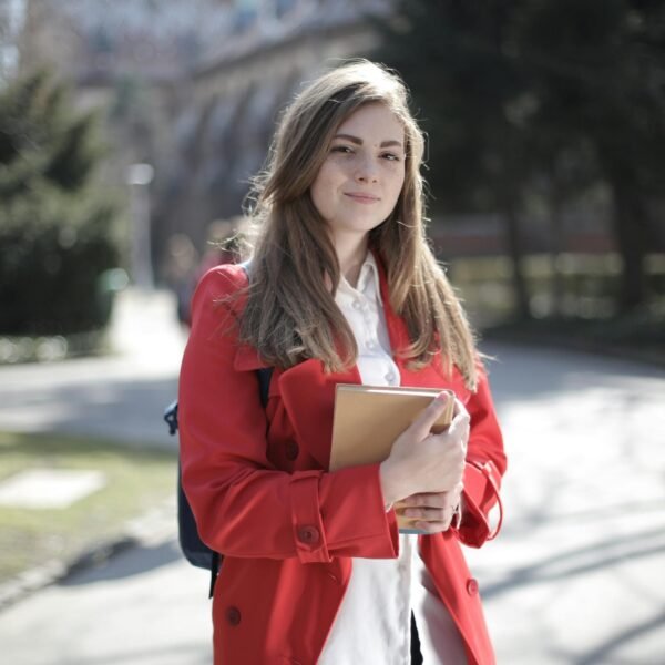 Young woman in a red coat smiling while holding a book outside on a sunny day.