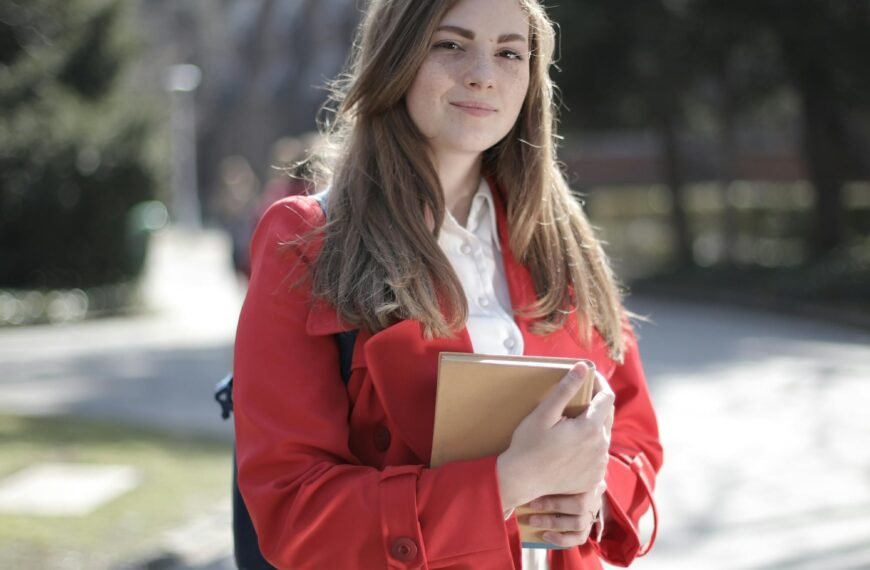 Young woman in a red coat smiling while holding a book outside on a sunny day.