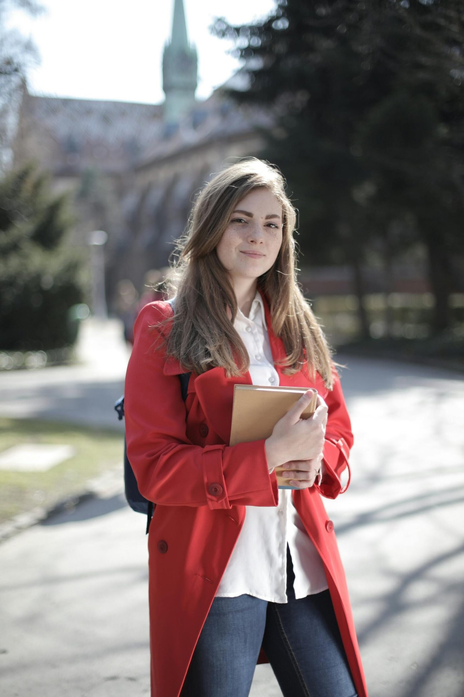 Young woman in a red coat smiling while holding a book outside on a sunny day.