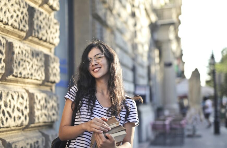 Happy young woman standing outdoors holding books, smiling in city setting.