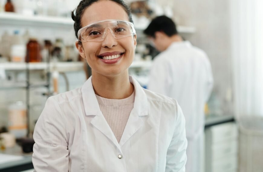 A confident female scientist in a lab coat smiling, working in an indoor laboratory environment.