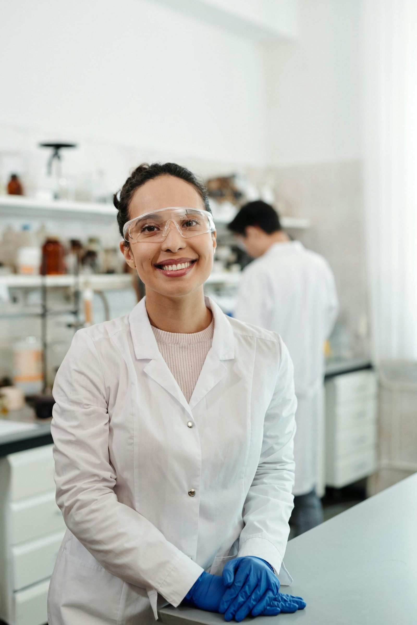 A confident female scientist in a lab coat smiling, working in an indoor laboratory environment.