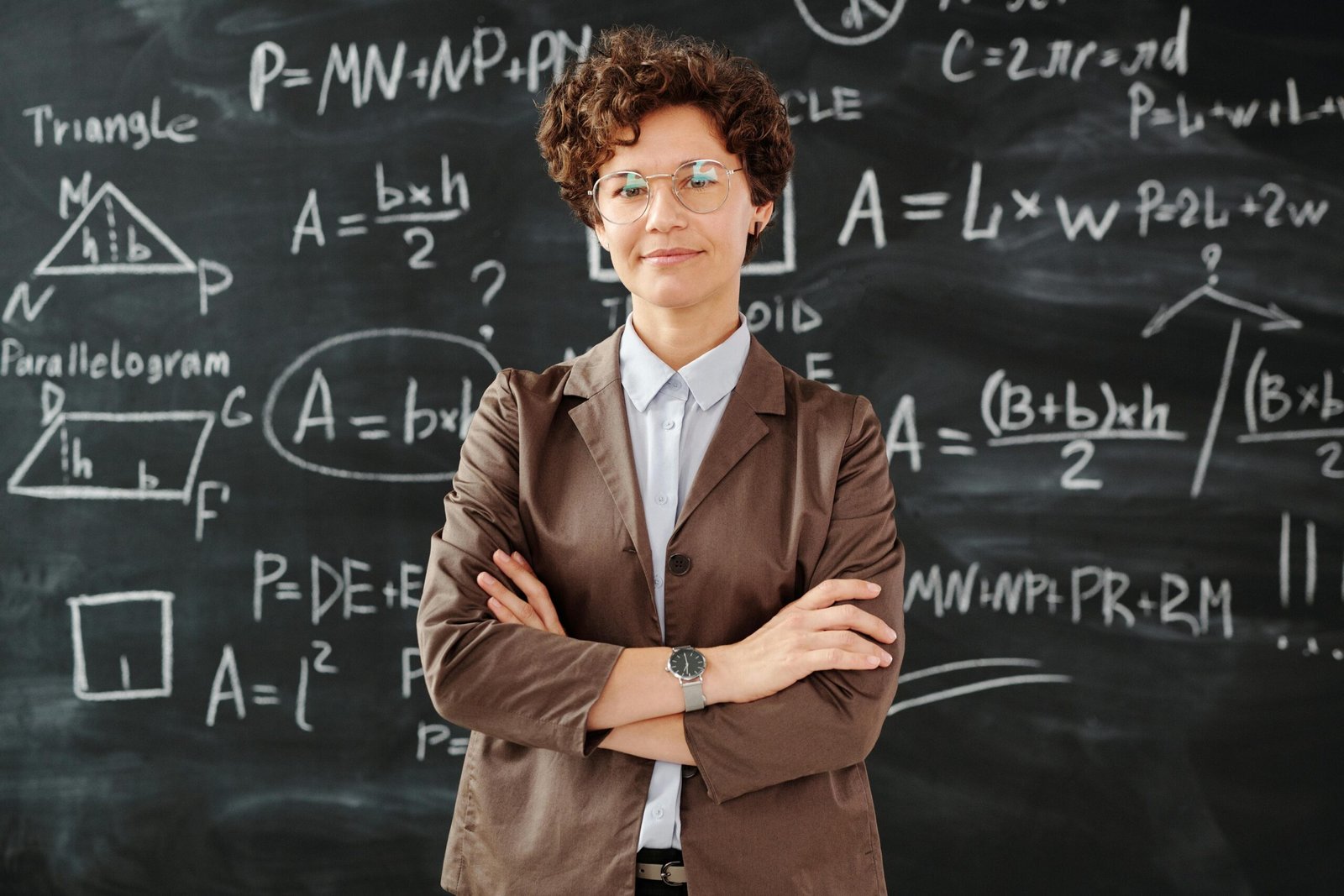 Confident female teacher standing with arms crossed in front of a detailed mathematical blackboard.