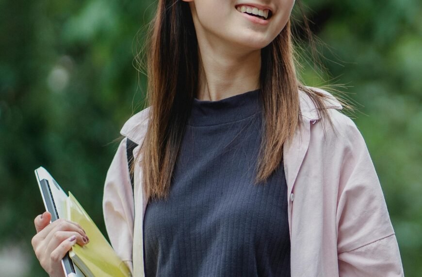 Smiling young Asian woman carrying a laptop and books, enjoying a sunny day outdoors.