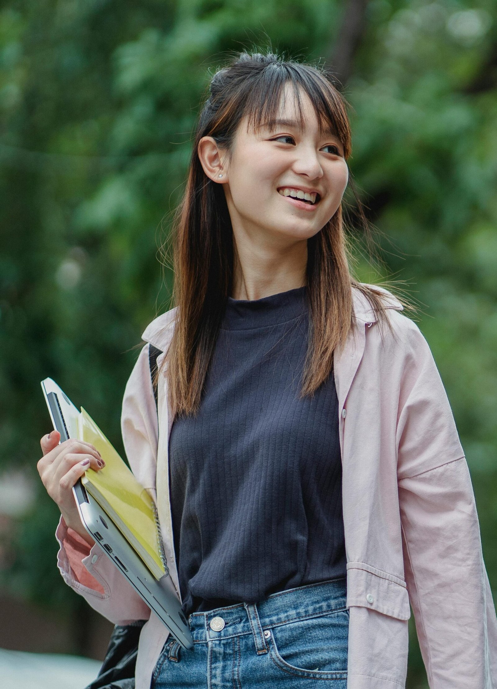 Smiling young Asian woman carrying a laptop and books, enjoying a sunny day outdoors.