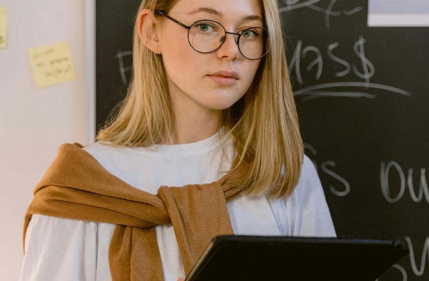 Caucasian woman with eyeglasses holds tablet in a classroom, surrounded by sticky notes and blackboard.