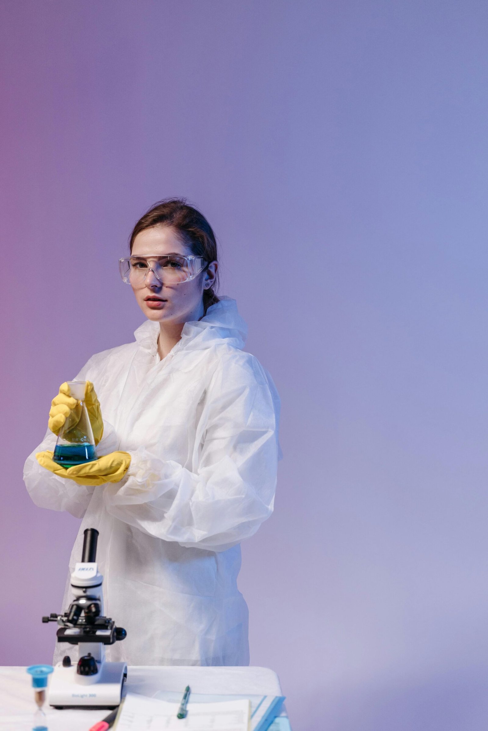 A woman scientist in lab gear analyzes a sample beside a microscope in a lab setting.