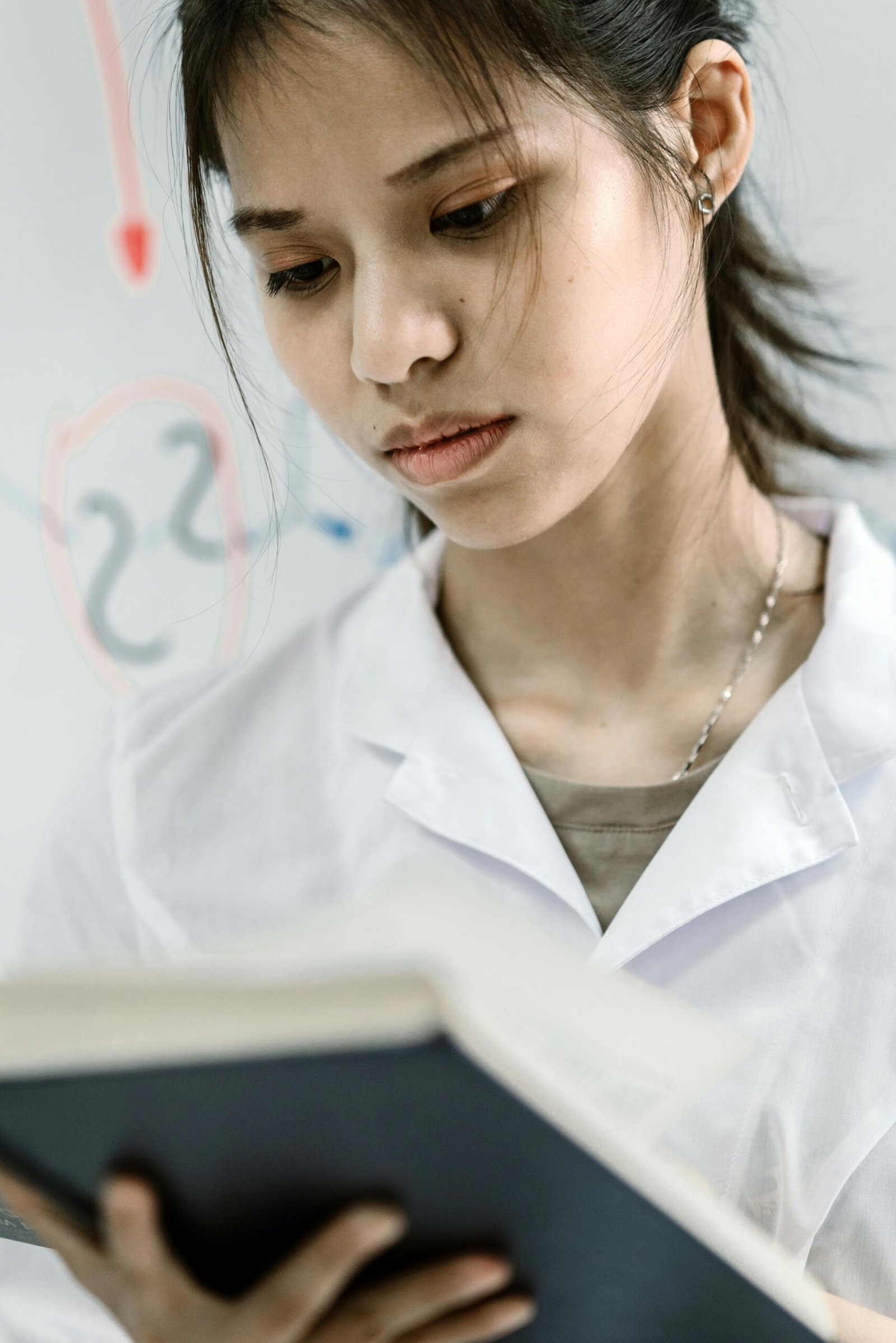 Asian female scientist in lab coat reading a book, experimenting in a laboratory setting.